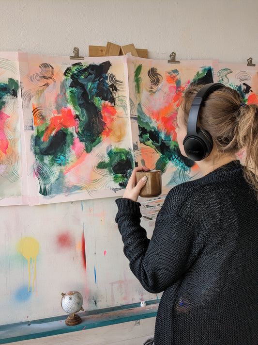 Woman artist with cup of favourite drink looking at work in progress abstract paintings with bold orange pink colors and deep greens and blues